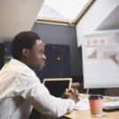 Attractive young African CEO sitting in conference room during meeting developing strategy, beautiful female manager presenting project on flip chart in background, explaining ideas and team goals